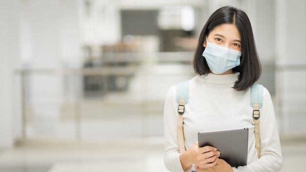 Retrato de un joven estudiante universitario adolescente asiático con vestimenta informal, sostiene una tableta con una mochila escolar, usa una mascarilla médica y mantiene el distanciamiento social durante la pandemia de COVID-19 en el campus universitario