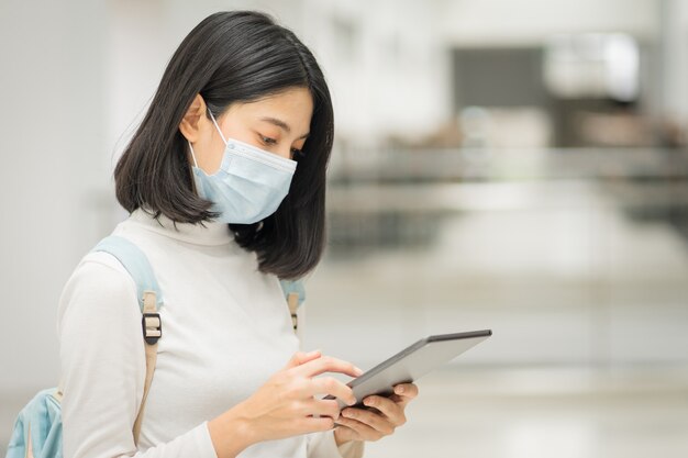 Retrato de un joven estudiante universitario adolescente asiático con vestimenta informal, sostiene una tableta con una mochila escolar, usa una mascarilla médica y mantiene el distanciamiento social durante la pandemia de COVID-19 en el campus universitario