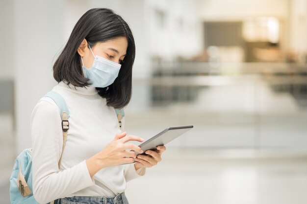 Retrato de un joven estudiante universitario adolescente asiático con vestimenta informal, sostiene una tableta con una mochila escolar, usa una mascarilla médica y mantiene el distanciamiento social durante la pandemia de COVID-19 en el campus universitario