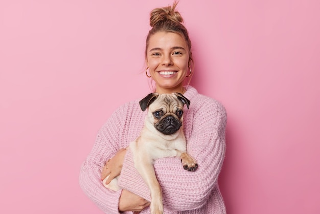 Foto retrato de una joven europea feliz que lleva un perro pug expresa amor y cuidado a su mascota favorita sonríe alegremente aislada sobre un fondo rosado contenta de tener un nuevo miembro de la familia concepto de animales