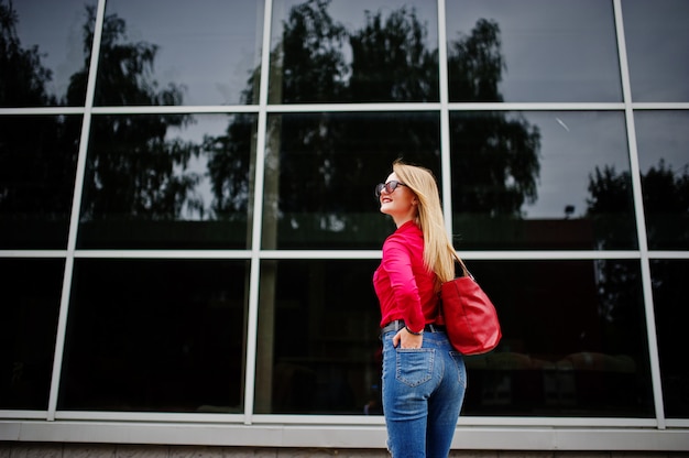 Retrato de una joven fabulosa en blusa roja y jeans posando con su bolso y gafas de sol fuera del centro comercial