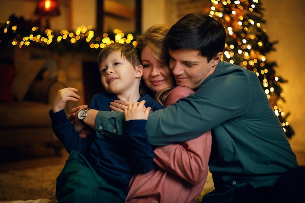 Foto retrato de joven familia caucásica abrazando con su hijo posando en el piso en el acogedor interior de navidad