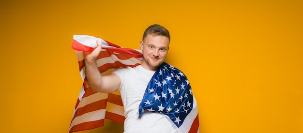 Retrato de un joven feliz con barba en ropa casual sosteniendo la bandera estadounidense en un amarillo