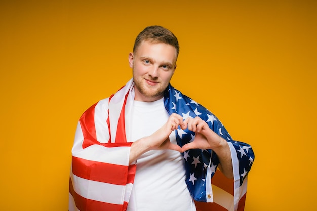 Retrato de un joven feliz con barba en ropa casual sosteniendo la bandera estadounidense en un amarillo