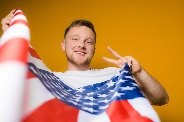 Retrato de un joven feliz con barba en ropa casual sosteniendo la bandera estadounidense en un amarillo