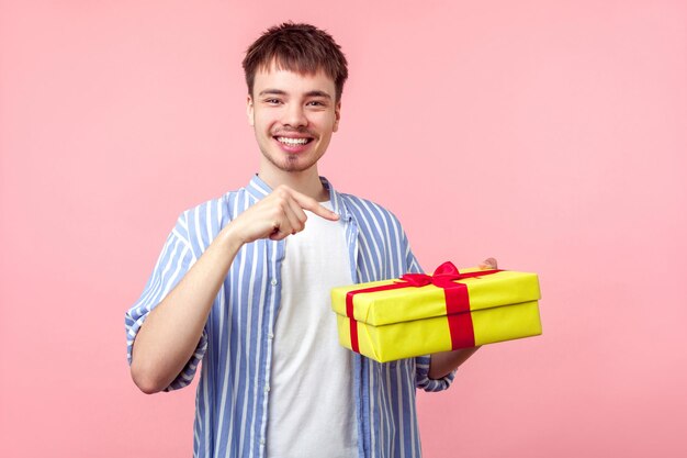 Retrato de un joven feliz de cabello castaño con barba pequeña y bigote en pantalones casual apuntando a una caja de regalo y sonriendo a la cámara disfrutando de una foto de estudio interior aislada en un fondo rosado