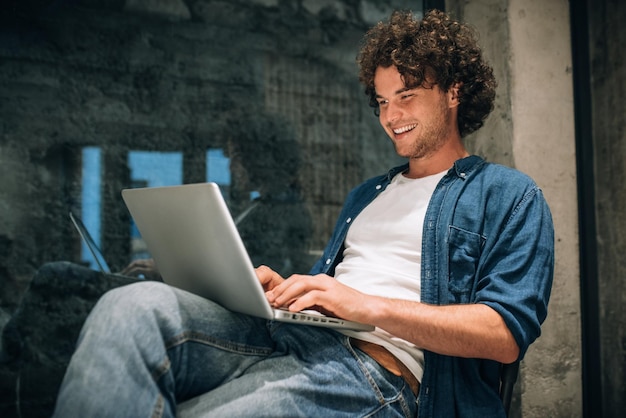 Foto retrato de un joven feliz con cabello rizado usando una computadora portátil para trabajar en línea hombre independiente inteligente chateando en línea con clientes hombre guapo leyendo noticias escribiendo en su computadora portátil en la oficina