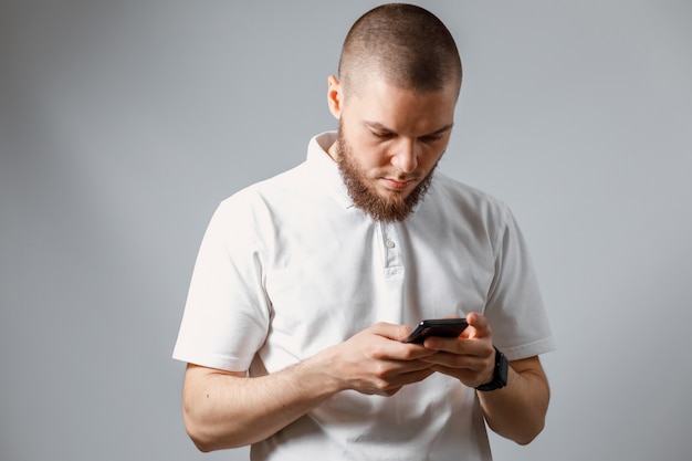 Retrato de un joven feliz en una camiseta blanca mirando cuidadosamente el teléfono en gris.