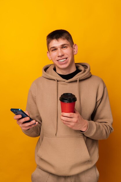 Foto retrato de un joven feliz con capucha sosteniendo un teléfono inteligente y una taza de café para llevar