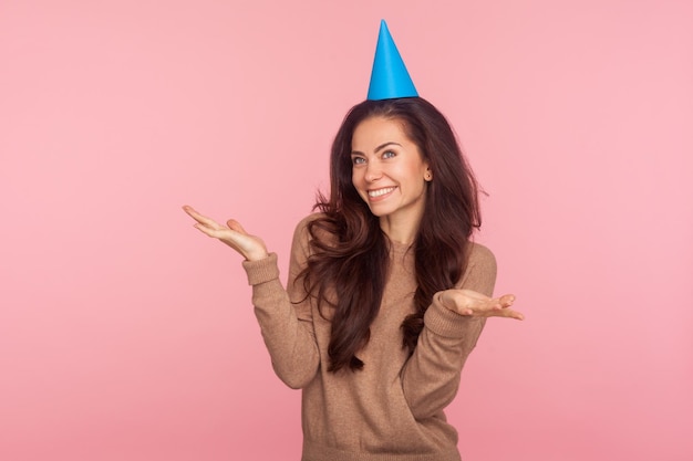 Retrato de una joven feliz y encantada con el pelo ondulado moreno usando un cono divertido y levantando las manos sorprendida y sorprendida por la fiesta de cumpleaños en un estudio interior aislado en un fondo rosa