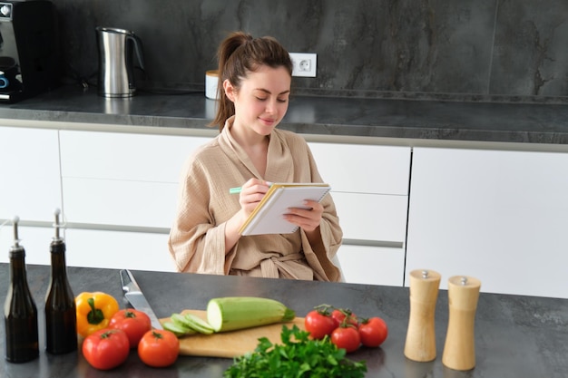 Retrato de una joven feliz escribe el menú para la cena se sienta en la cocina cerca de las verduras