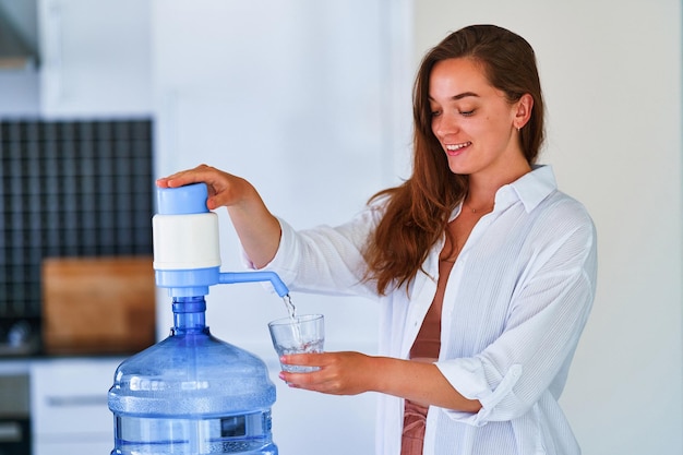 Foto retrato de una joven feliz y linda mujer sonriente que vierte agua limpia y fresca en un vaso de una botella grande en casa