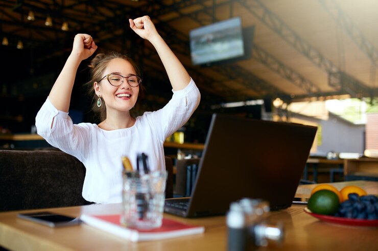 Foto retrato de una joven feliz mujer de negocios celebrando el éxito con los brazos levantados frente a la computadora portátil mujer de raza mixta ganó mucho dinero en el premio de la lotería brazos levantados con los puños freelancer terminó el proyecto