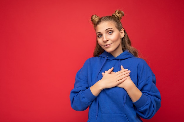 Retrato de joven feliz positiva hermosa mujer rubia con dos cuernos con emociones sinceras vistiendo casual sudadera con capucha azul brillante aislado sobre fondo rojo con espacio vacío y tomados de la mano en el corazón.