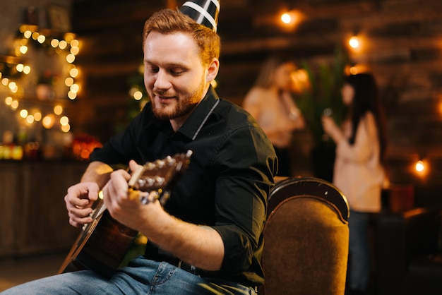 Retrato de joven feliz en el sombrero de fiesta tocando la guitarra en el contexto de hablar amigos. Árbol de Navidad con guirnalda y pared con iluminación festiva de fondo.