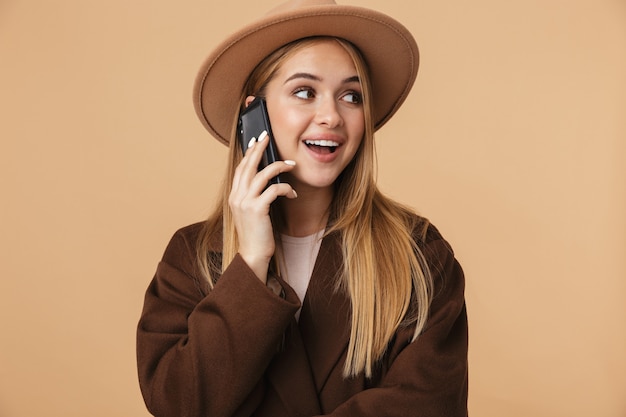 Retrato de joven feliz con sombrero sonriendo y hablando por teléfono celular aislado en beige