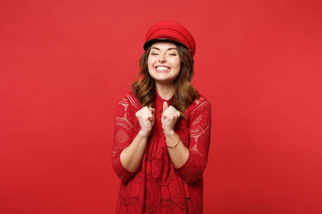 Retrato de una joven feliz y sonriente con vestido de encaje y puños apretados con gorra aislada en un fondo de pared rojo brillante en el estudio. Emociones sinceras de la gente, concepto de estilo de vida. Simulacros de espacio de copia.