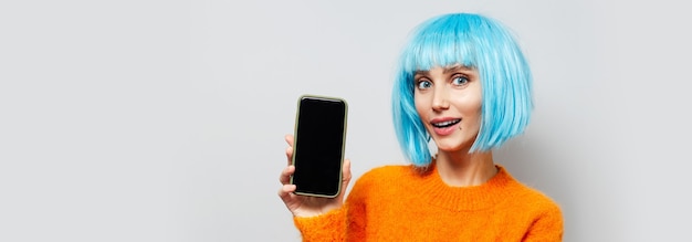 Retrato de joven feliz sosteniendo smartphone en mano, vistiendo peluca de pelo azul y suéter naranja