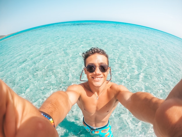 Retrato de joven feliz tomando un selfie de él en la playa en un agua azul turquesa divirtiéndose y disfrutando de unas vacaciones solos al aire libre.