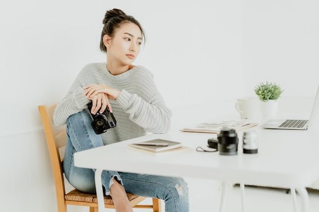 Retrato de una joven fotógrafa asiática con una cámara profesional sentada en una silla y mirando hacia otro lado mientras trabajaba en su estudio Startup independiente Propietario de una pequeña empresa Marketing en línea PYME