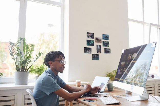 Retrato de joven fotógrafo afroamericano con computadora en el escritorio en la oficina en casa con software de edición de fotos en pantalla, espacio de copia