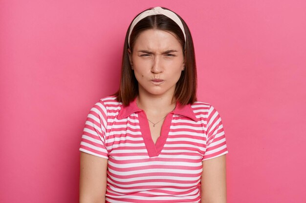 Retrato de una joven frustrada y enfadada con el pelo castaño y una camiseta informal a rayas de pie aislada con un fondo rosa, dientes apretados y ojos cerrados discutiendo expresando emociones negativas y odio