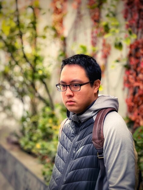 Retrato de un joven con gafas de pie al aire libre contra la hiedra roja en la pared y las plantas