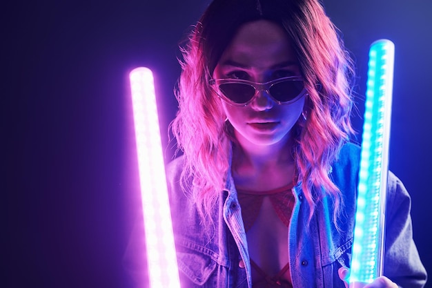 Retrato de una joven con gafas sosteniendo palos de luz en neón rojo y azul en el estudio