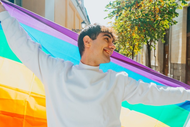 Retrato de joven gay con bandera lgtbi lgbtq en la ciudad en un día de verano entre los edificios, caminando sonriendo luchando contra la discriminación. Vista frontal. Celebrando el orgullo con espacio de copia