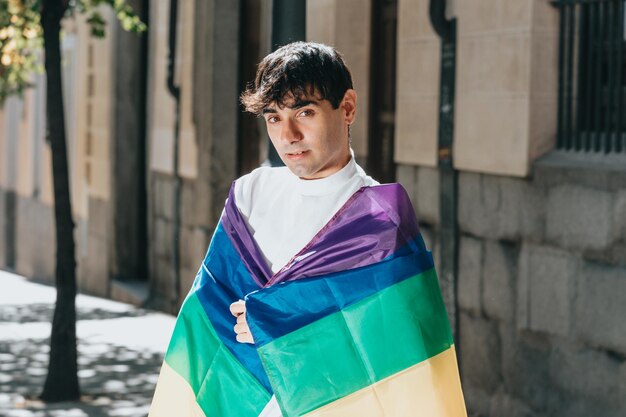 Foto retrato de joven gay con bandera lgtbi lgbtq en la ciudad en un día de verano entre los edificios, luchando seriamente contra la discriminación. vista frontal. celebrando el orgullo con espacio de copia