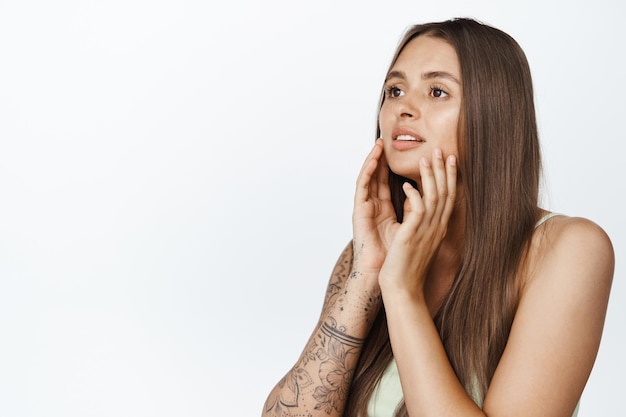 Retrato de una joven glamurosa con piel y cabello sanos, tocándose la cara con maquillaje, mirando a un lado, de pie contra un fondo blanco