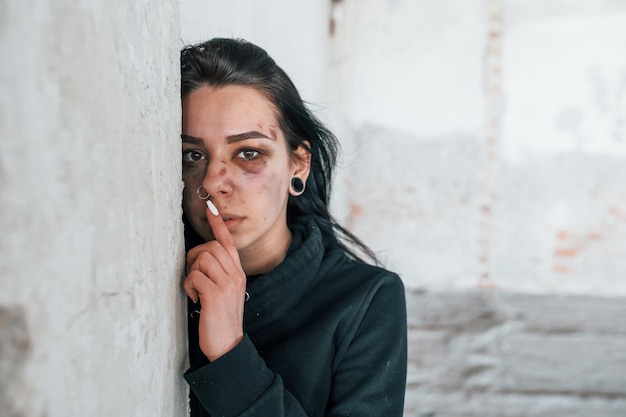 Retrato de una joven golpeada con moretones bajo los ojos en el interior de un edificio abandonado que muestra un gesto de silencio