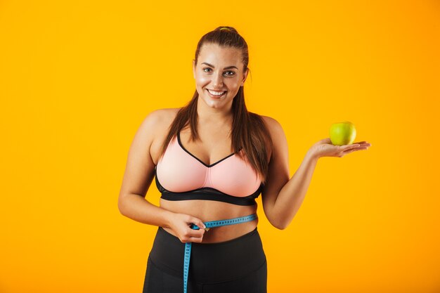 Retrato de joven gordita en sujetador deportivo midiendo su cintura con medidor y sosteniendo apple, aislado sobre fondo amarillo