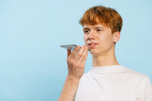 Retrato de un joven grabando un mensaje de voz en un teléfono aislado sobre fondo azul de estudio Comunicación