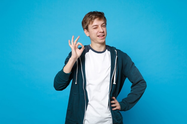 Retrato de joven gracioso parpadeante en ropa casual de pie y mostrando gesto OK aislado en la pared azul.