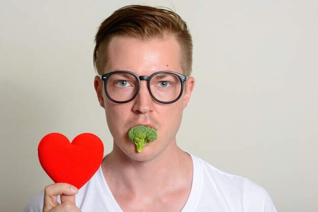 Retrato de joven guapo con anteojos comiendo brócoli y sosteniendo el corazón