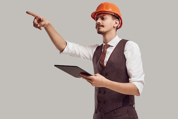 Retrato de joven y guapo arquitecto árabe confiado con bigote elegante en traje de lana y casco de construcción tiene tableta