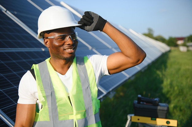 Retrato de un joven y guapo artesano afroamericano con casco protector Hombre en uniforme y con herramientas de pie entre paneles solares