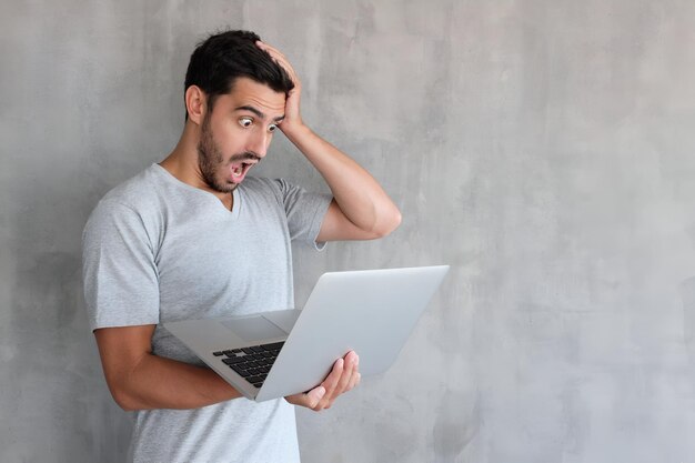 Foto retrato de un joven guapo con la boca abierta en una camiseta gris sosteniendo una computadora portátil plateada mirando sorprendido y confundido