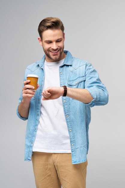 Retrato de joven guapo en camisa de jeans sobre pared ligera sosteniendo una taza de café para llevar