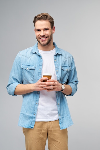 Retrato de joven guapo en camisa de jeans sobre pared ligera sosteniendo una taza de café para llevar