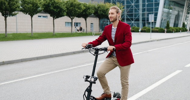 Retrato de joven guapo caucásico en chaqueta roja de pie al aire libre con bicicleta o scooter eléctrico y sonriendo a la cámara. Chico guapo con estilo en bicicleta en las calles de la ciudad.