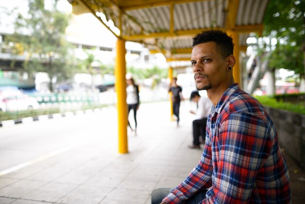 Retrato de joven guapo hipster africano barbudo esperando en la parada de autobús en la ciudad al aire libre