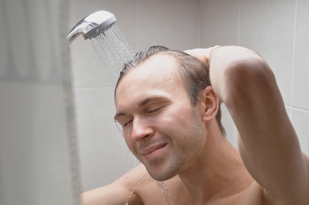 Retrato de joven guapo se lava con gel de ducha, hace espuma la cabeza con champú en el baño en casa de cerca