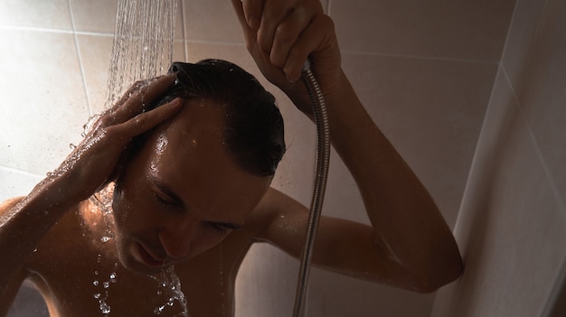 Retrato de joven guapo se lava con gel de ducha, hace espuma la cabeza con champú en el baño en casa de cerca