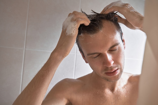 Retrato de joven guapo se lava con gel de ducha, hace espuma la cabeza con champú en el baño en casa de cerca