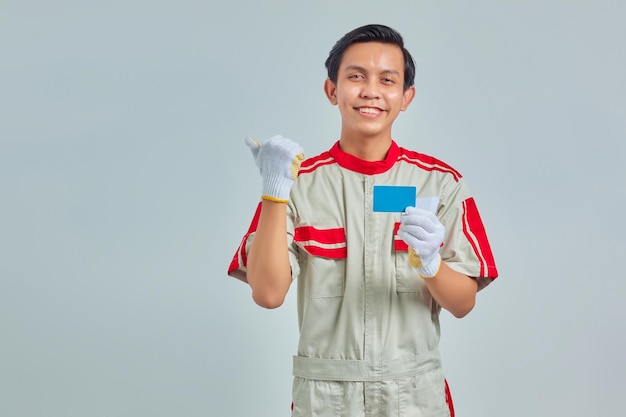 Retrato de joven guapo mecánico mostrando tarjeta de crédito y apuntando con el dedo al espacio vacío con expresión sonriente sobre fondo gris