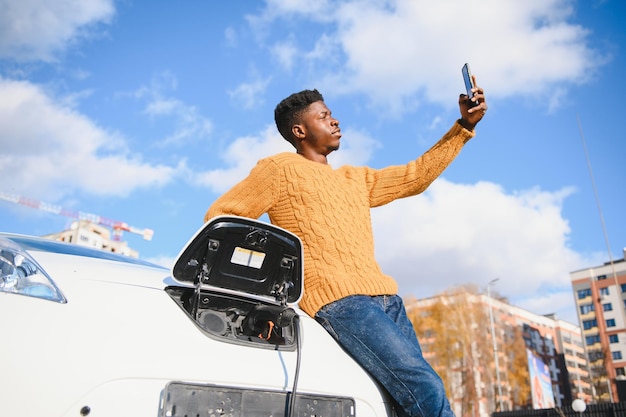 Retrato de joven guapo negro en traje de negocios, haciendo una foto selfie en su teléfono inteligente mientras se apoya en su automóvil eléctrico, cargando la batería en la central eléctrica de la ciudad