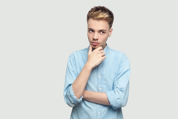 Retrato de joven guapo pensativo en camisa azul claro de pie, sosteniendo su barbilla, pensando y mirando a otro lado. tiro del estudio de interior en el espacio gris de la copia del fondo.