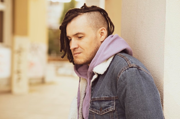 Retrato de joven guapo con rastas posando de pie apoyado contra la pared en la calle